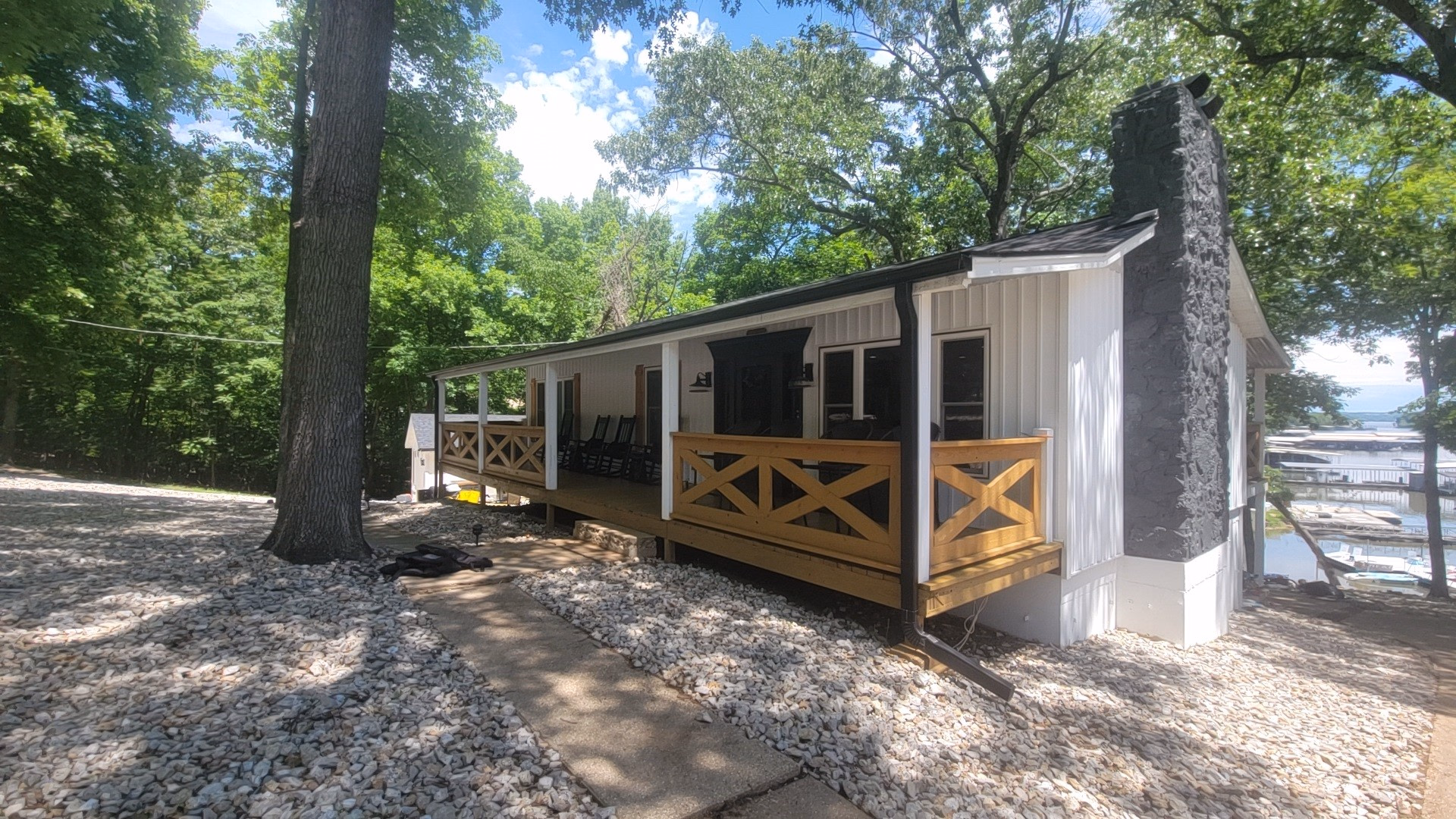 White cabin with wooden porch amid trees by a waterfront marina on a sunny day.