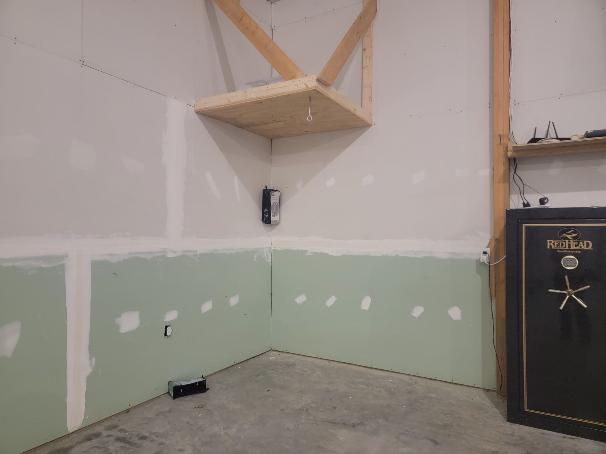 Corner of an unfinished room with green drywall, a wooden shelf above, and a black RedHead safe.