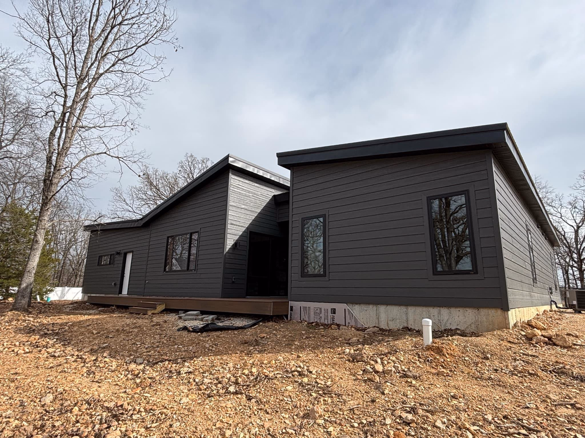 Modern dark gray house with angled roofs and multiple windows on uneven rocky soil.