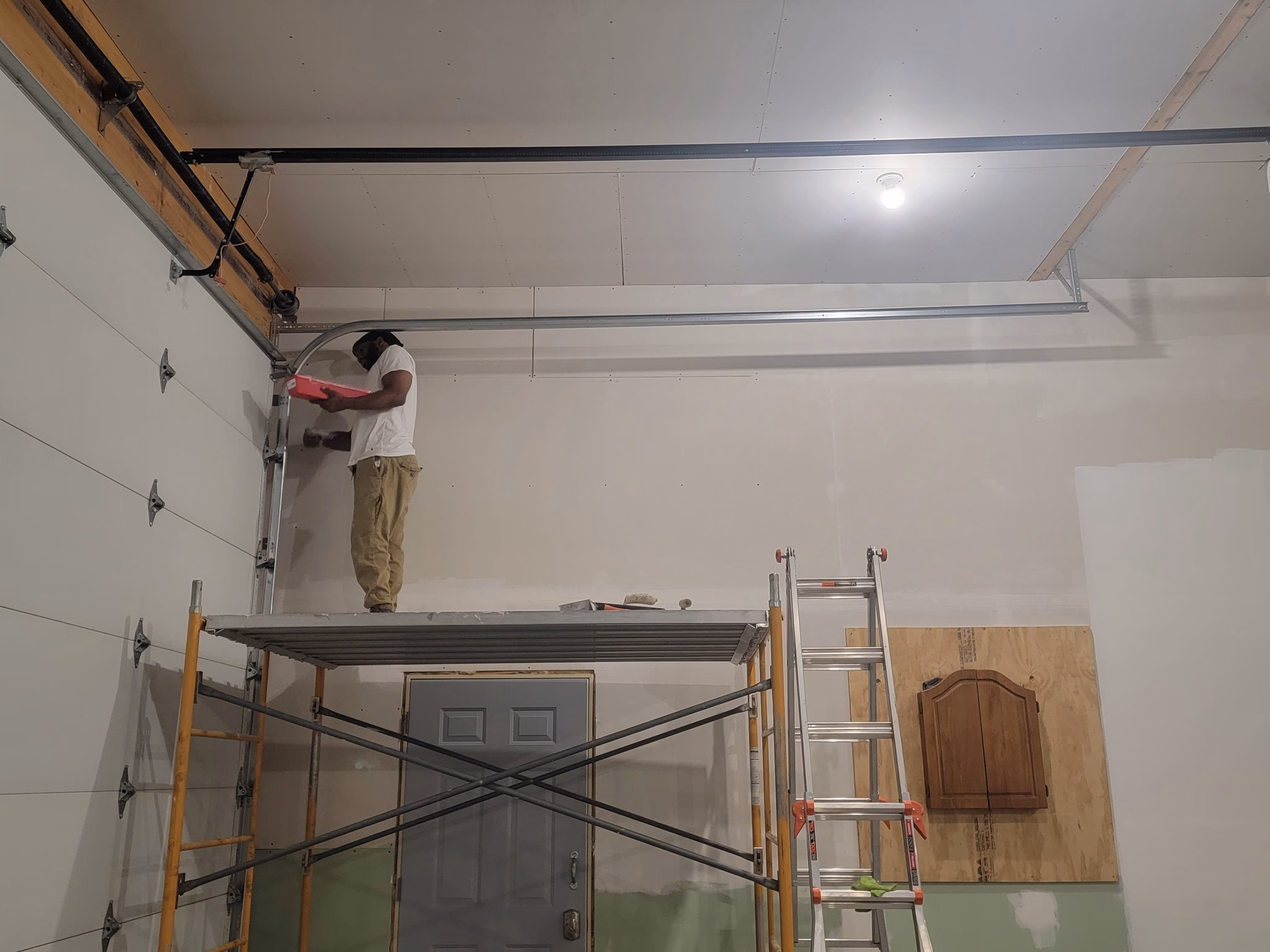 Worker on scaffolding applying drywall compound on a garage wall near a closed door and ladder.