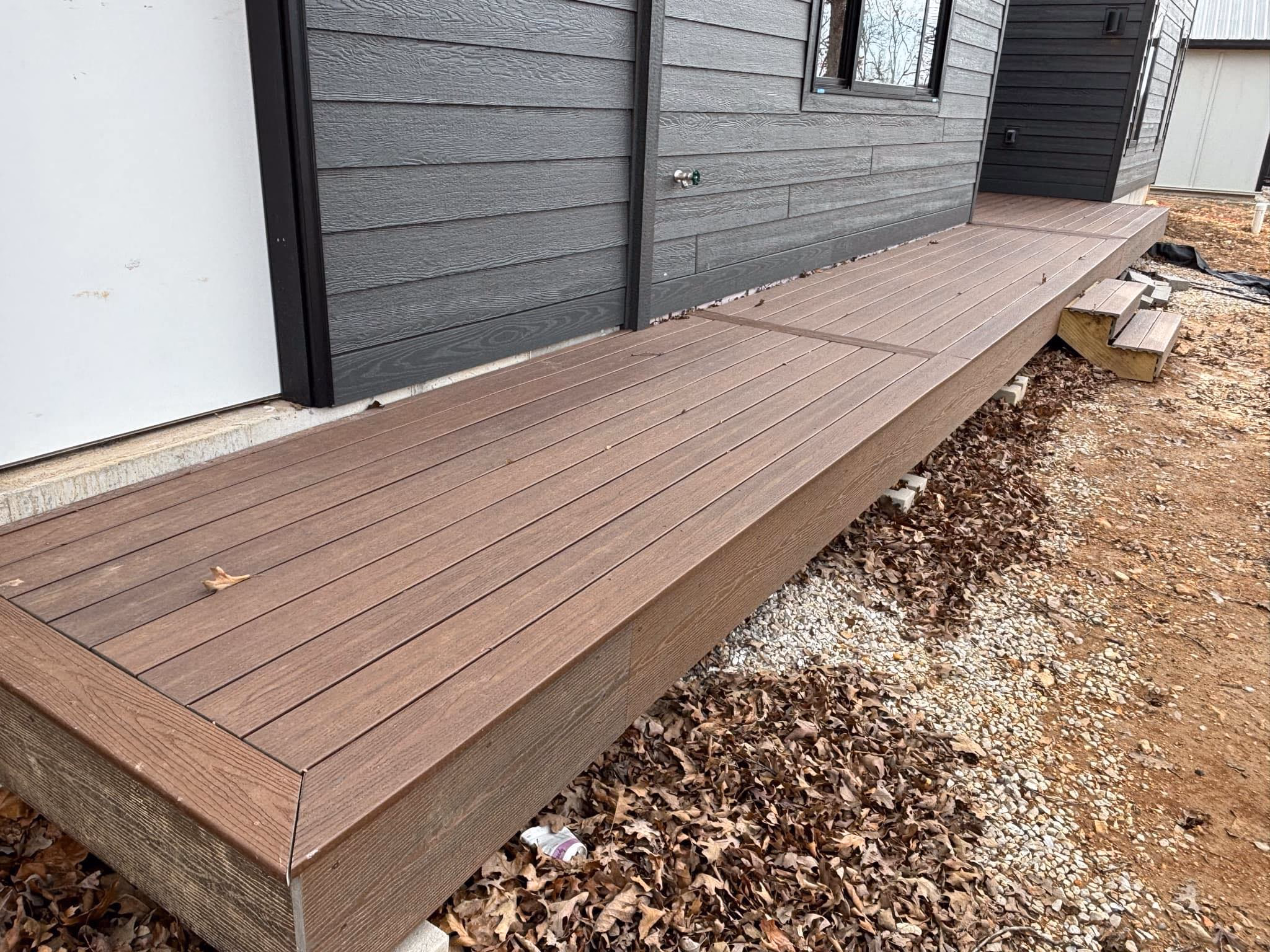 Brown wooden deck attached to dark gray house siding with small steps leading to dirt and leaves.