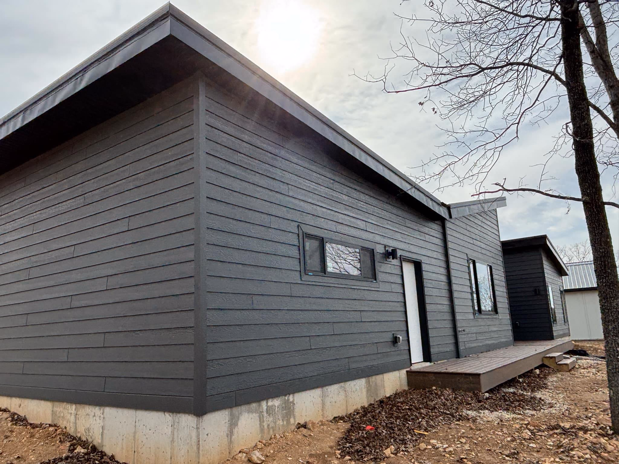 Modern dark gray house exterior with horizontal siding, white door, rectangular windows, and leaf-covered ground.