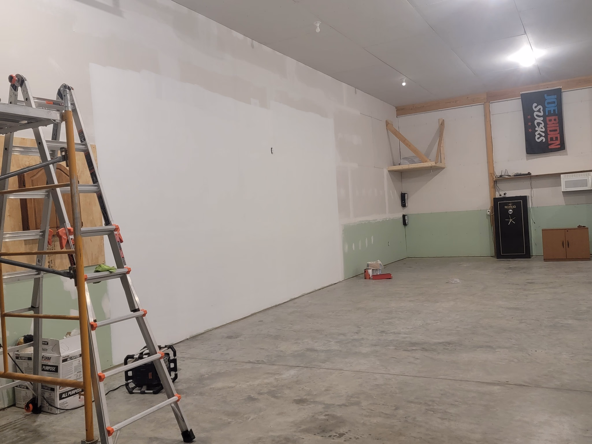 Partially painted room with ladder, boxes, wall safe, small shelf, and a banner reading Joe Biden Sucks.