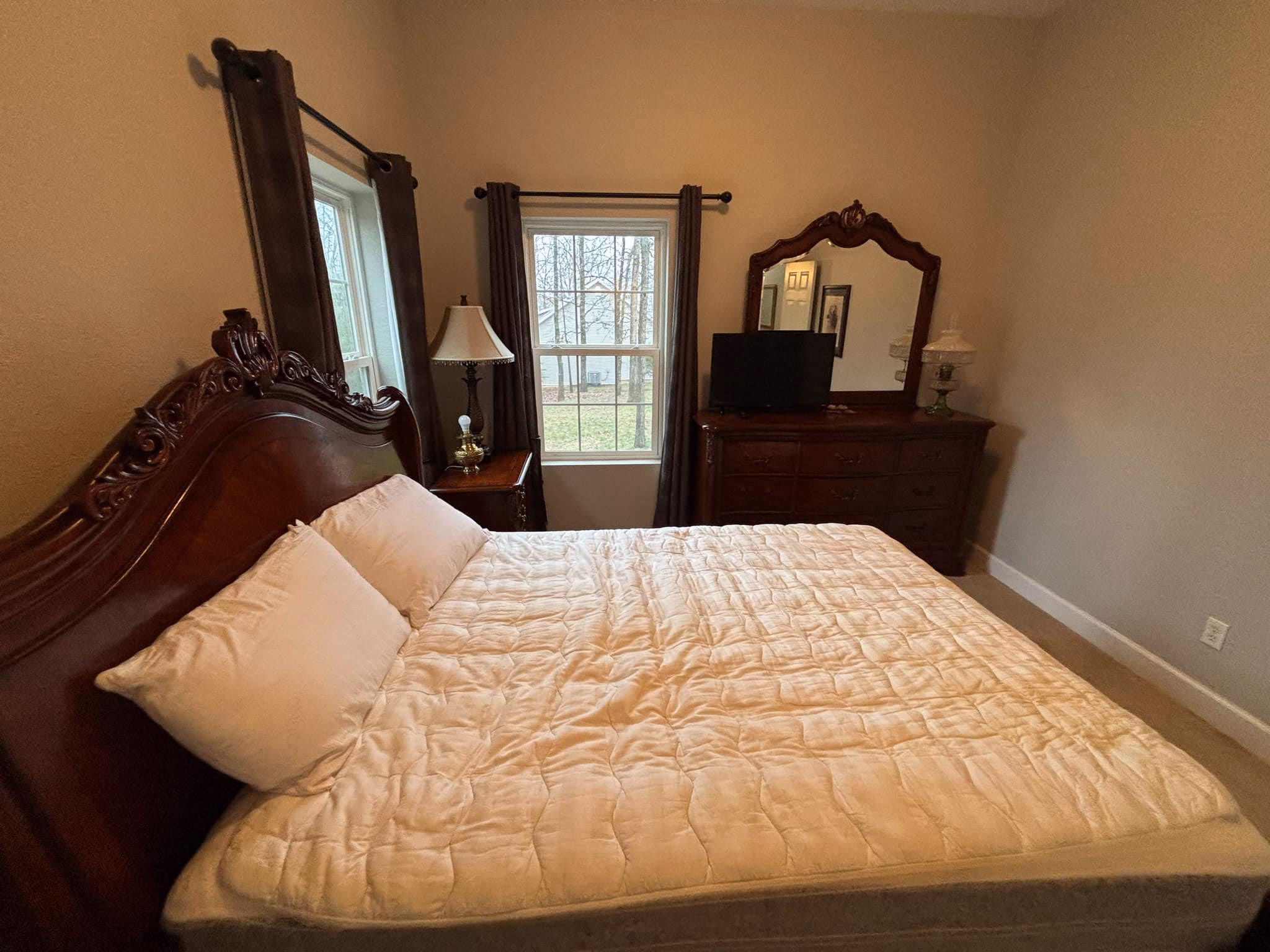 Bedroom with a carved wooden bed, beige comforter, two pillows, dresser with mirror, and two windows.