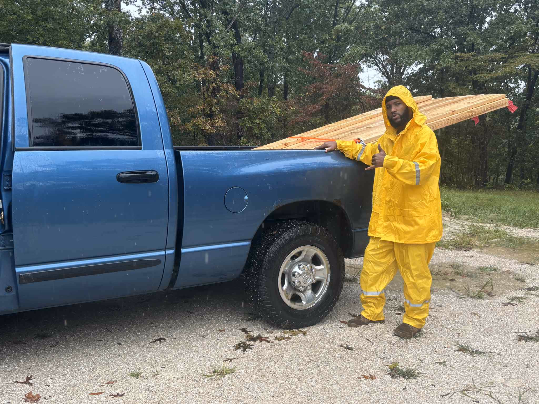 Man in yellow rain suit stands by blue pickup truck loaded with lumber on gravel road.
