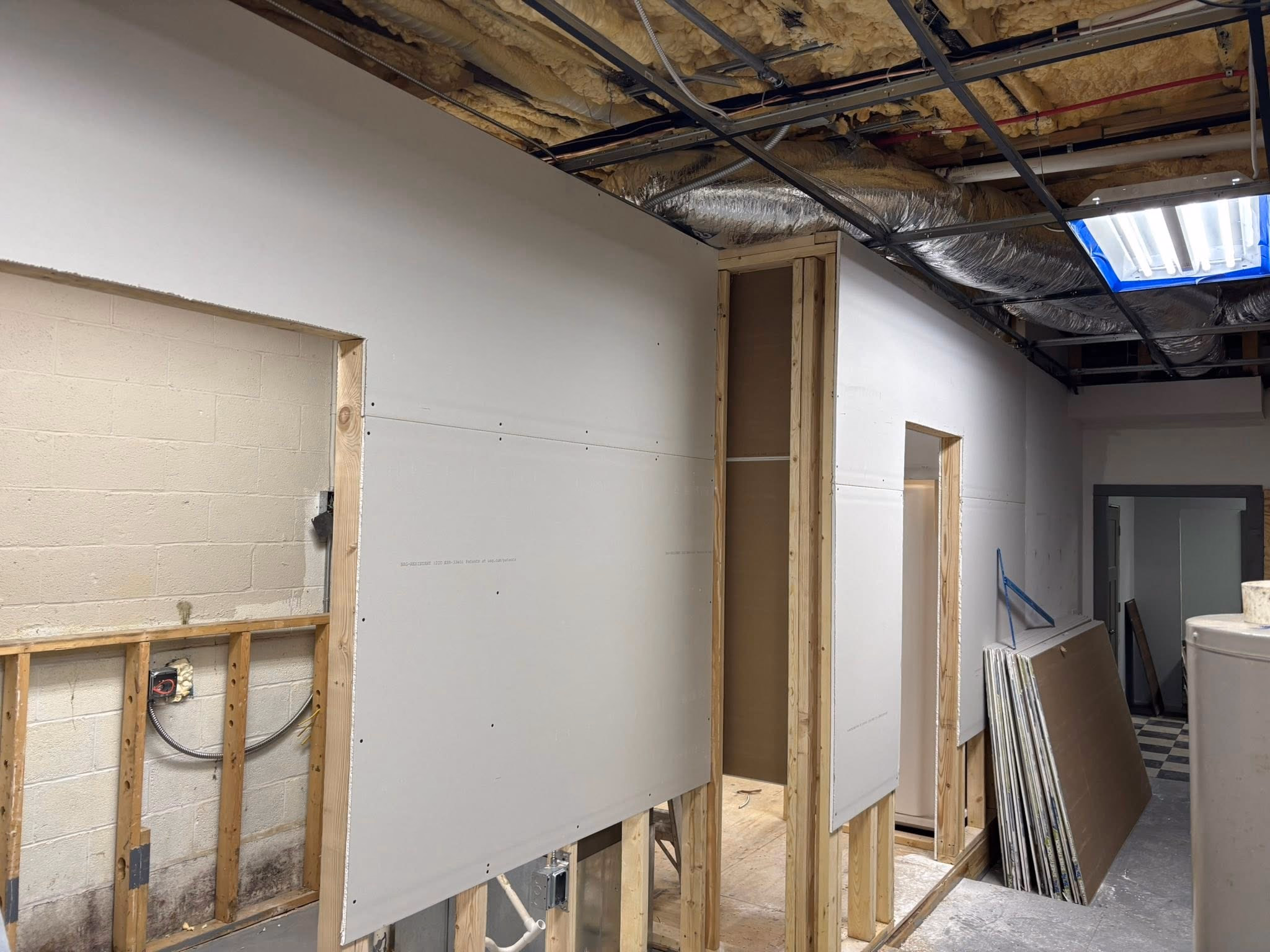 Interior construction site shows drywall installation and exposed ceiling insulation and ducts.