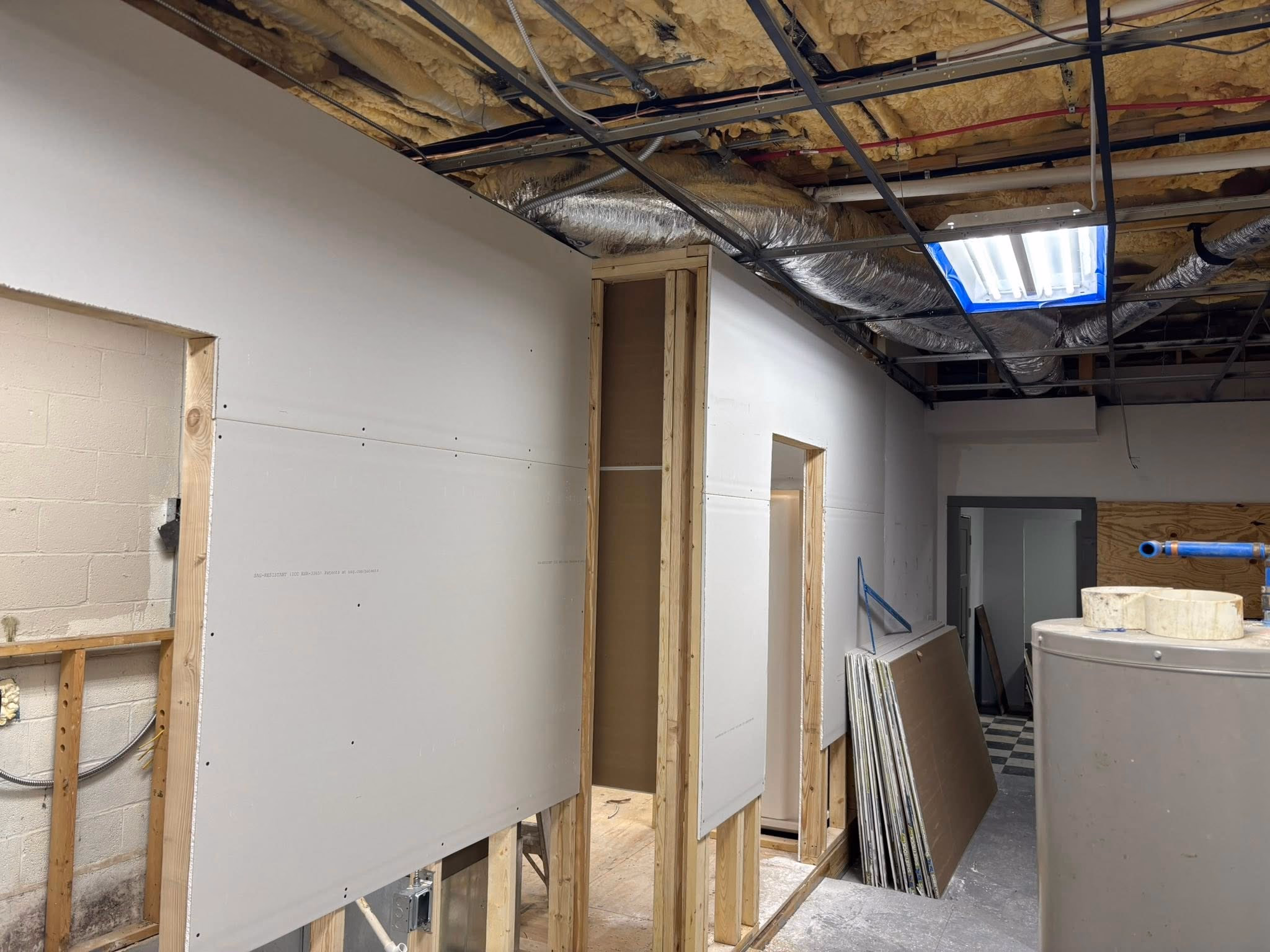 Interior construction site showing unfinished drywall walls, exposed ceiling insulation, and construction materials.