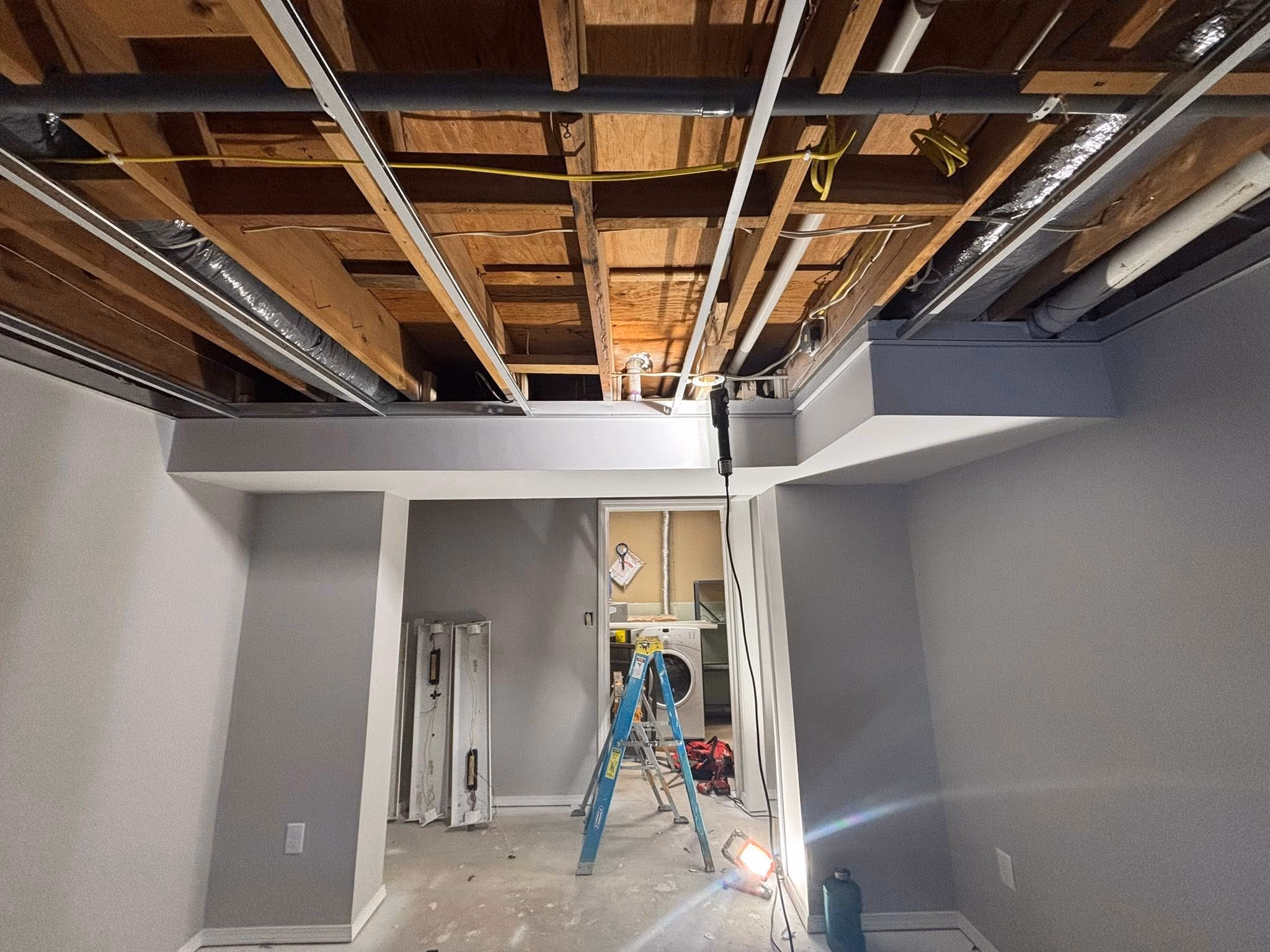 Basement under renovation with exposed ceiling, ladder, construction light, and washer-dryer in the back room.