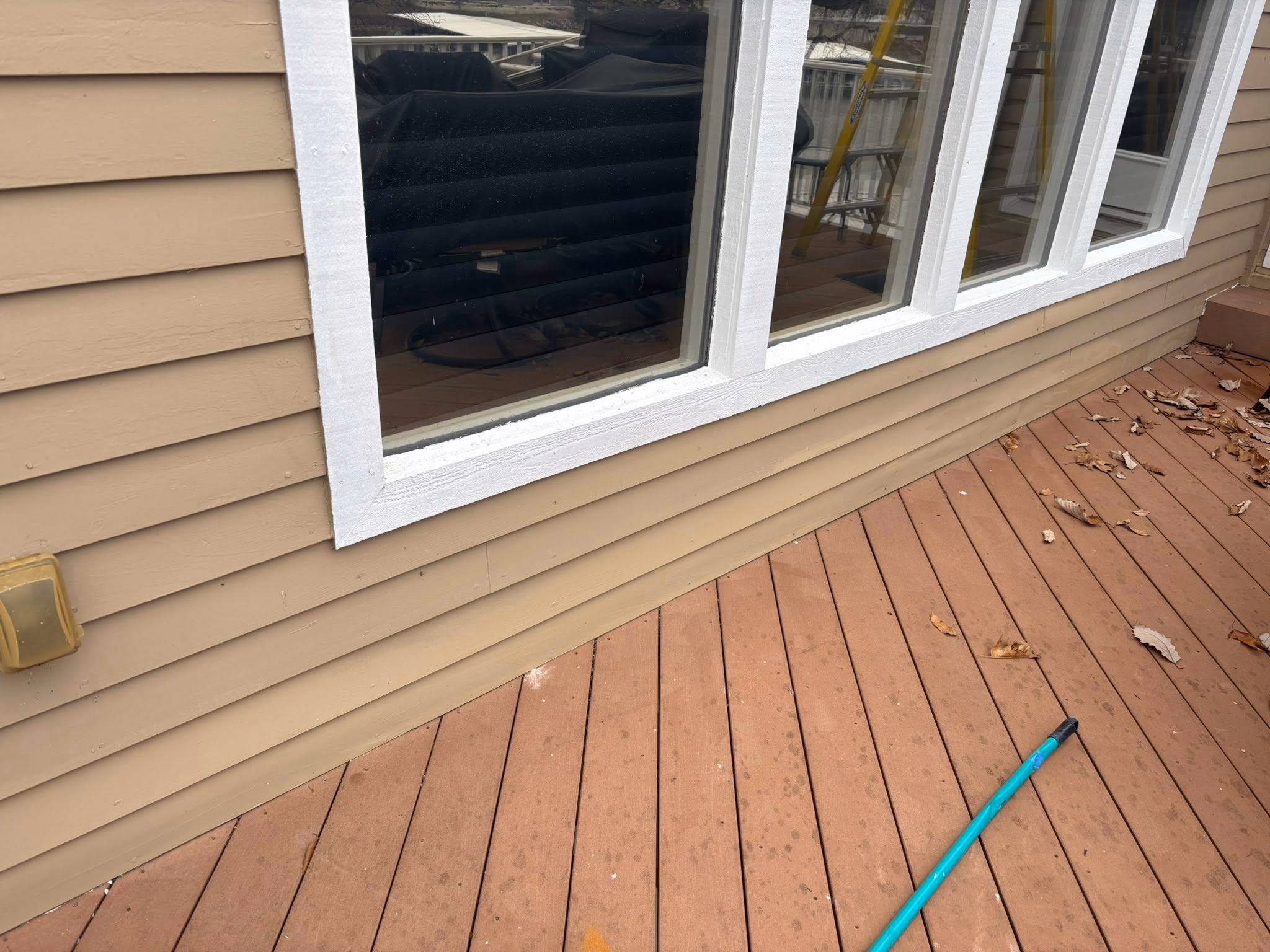 Beige siding walls with white window frames reflecting furniture and a brown wooden deck with scattered leaves and a broom.