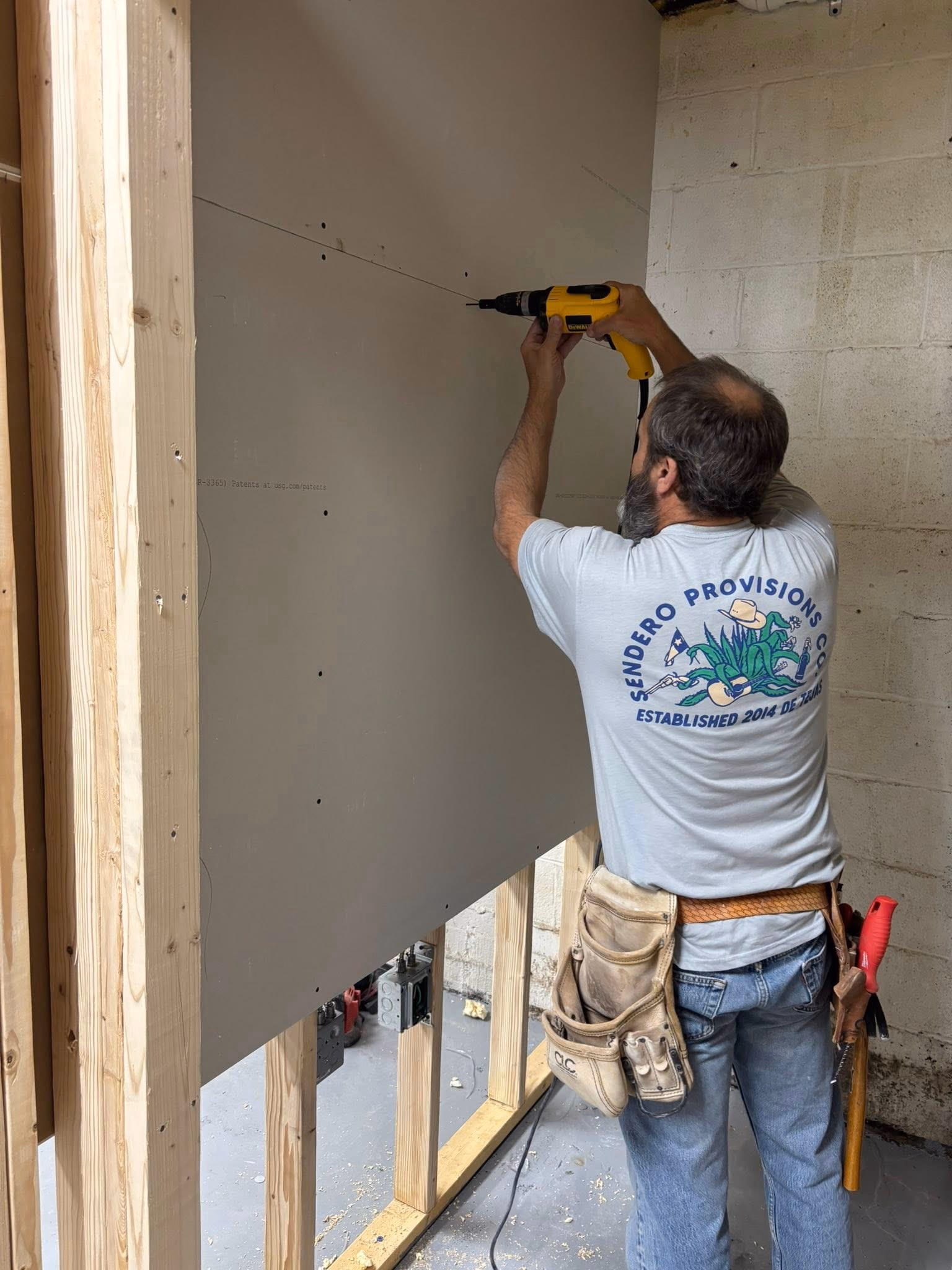 Man with tool belt using a yellow drill to install drywall on wooden studs in a construction area.