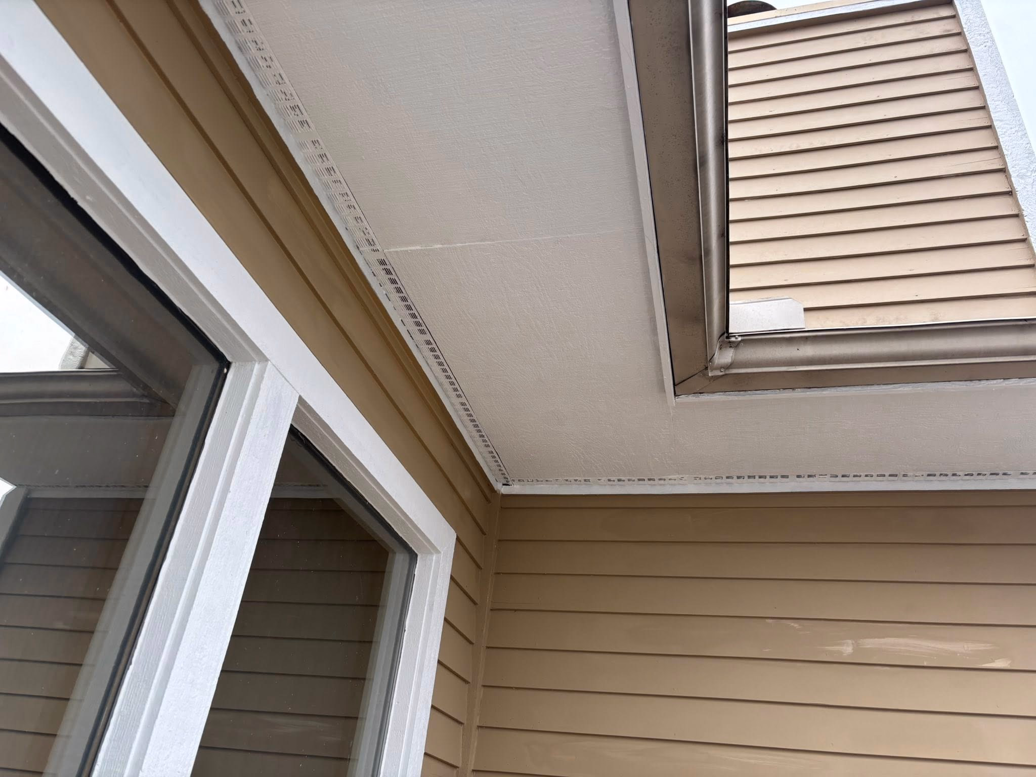 Beige siding on house exterior with white window frame and soffit vents along the ceiling.