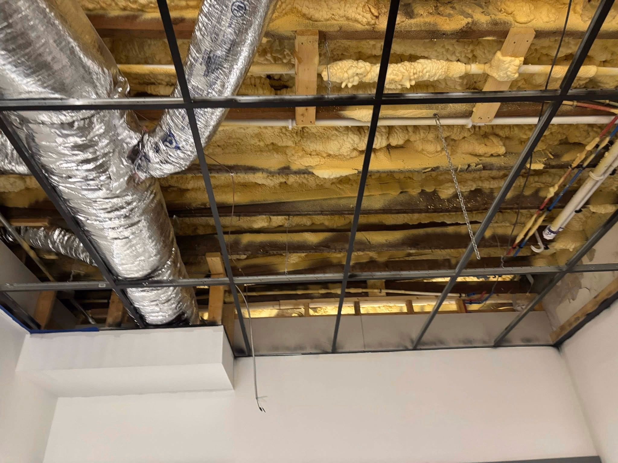 Ceiling under construction with exposed insulation, ductwork, pipes, and metal framing grid.