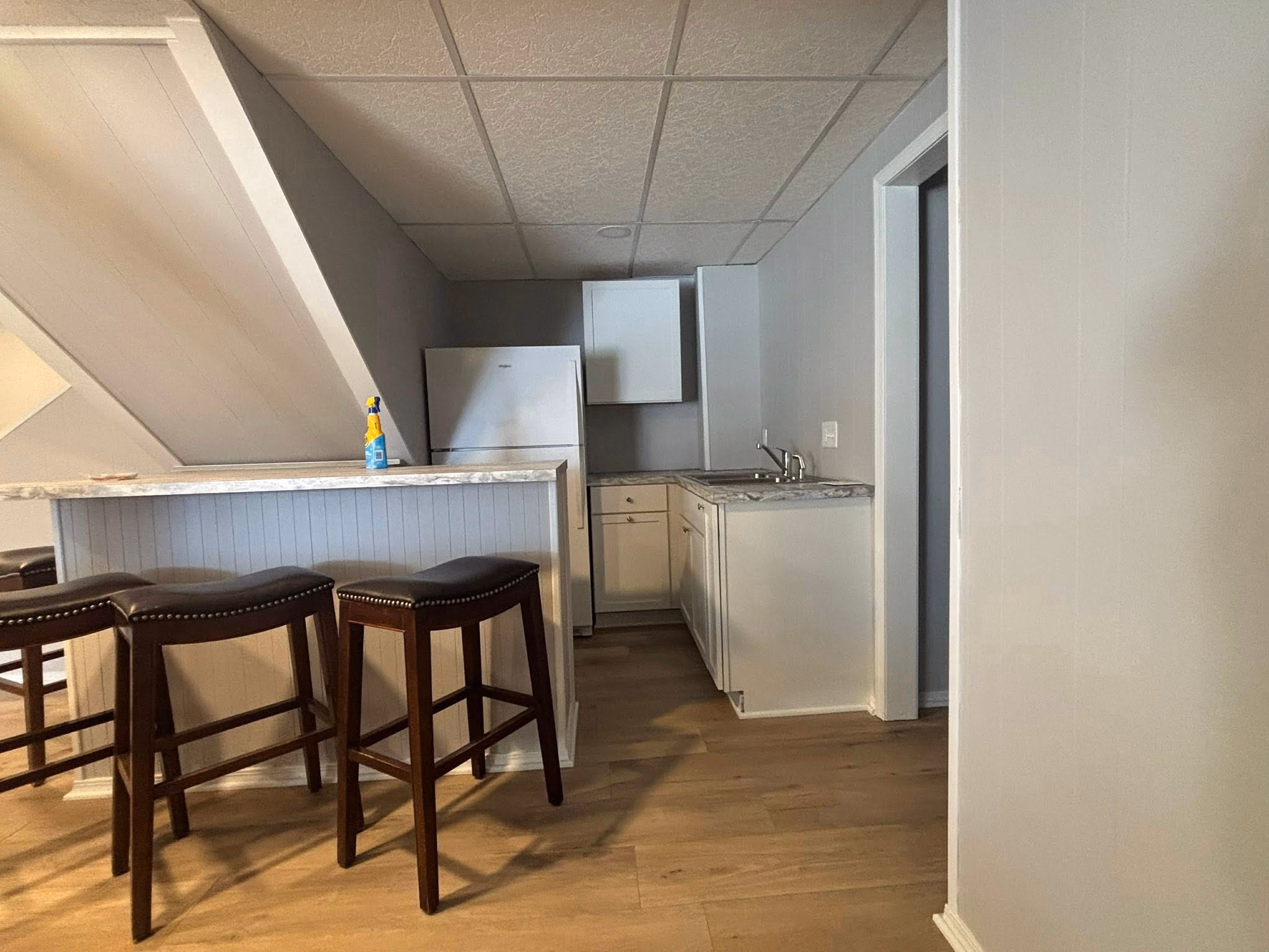 Small kitchen with white cabinets, refrigerator, sink, marble countertop, and two dark stools at a counter.