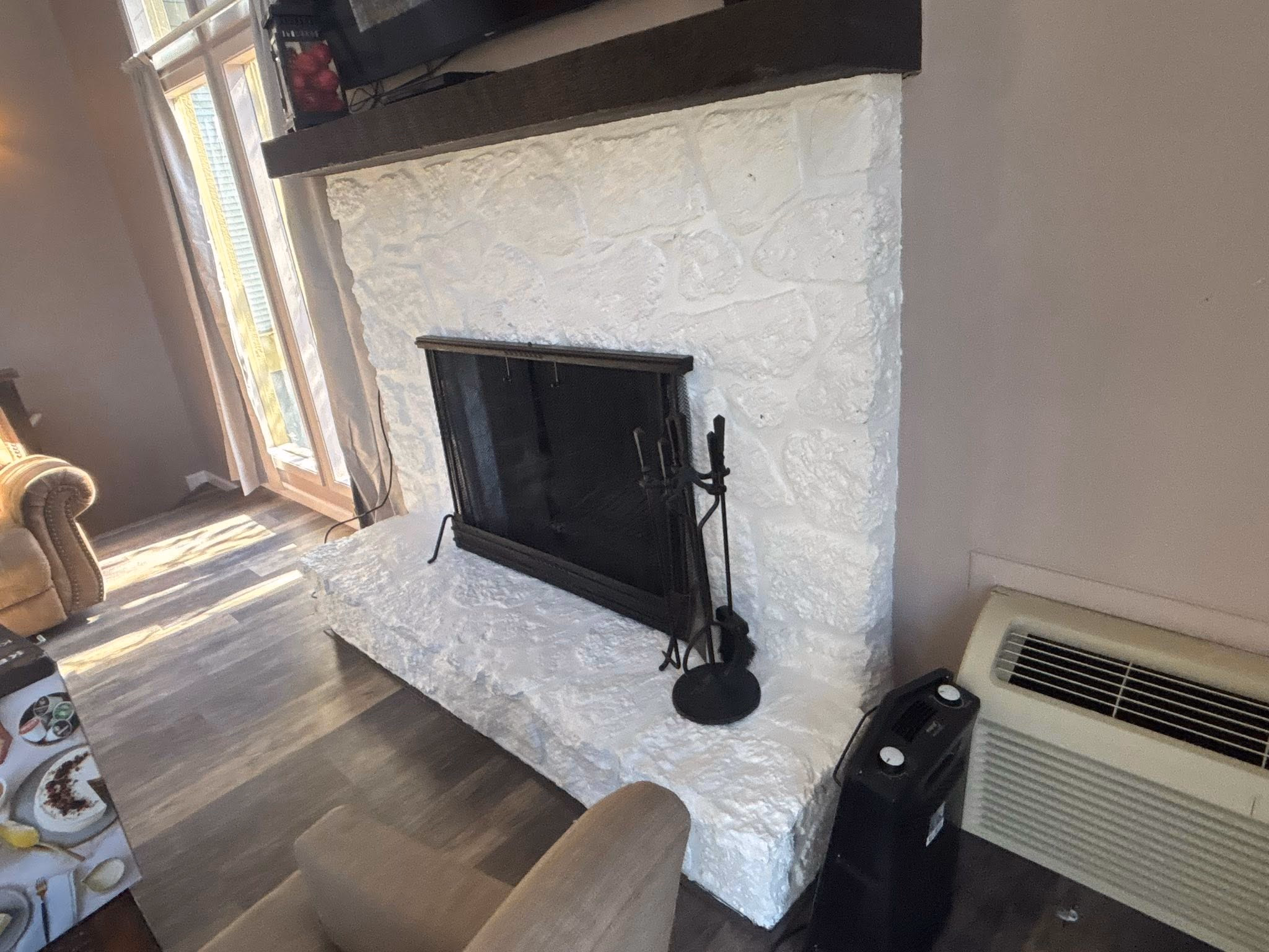 White stone fireplace with black screen and fire tools, next to an air conditioning unit and armchair.