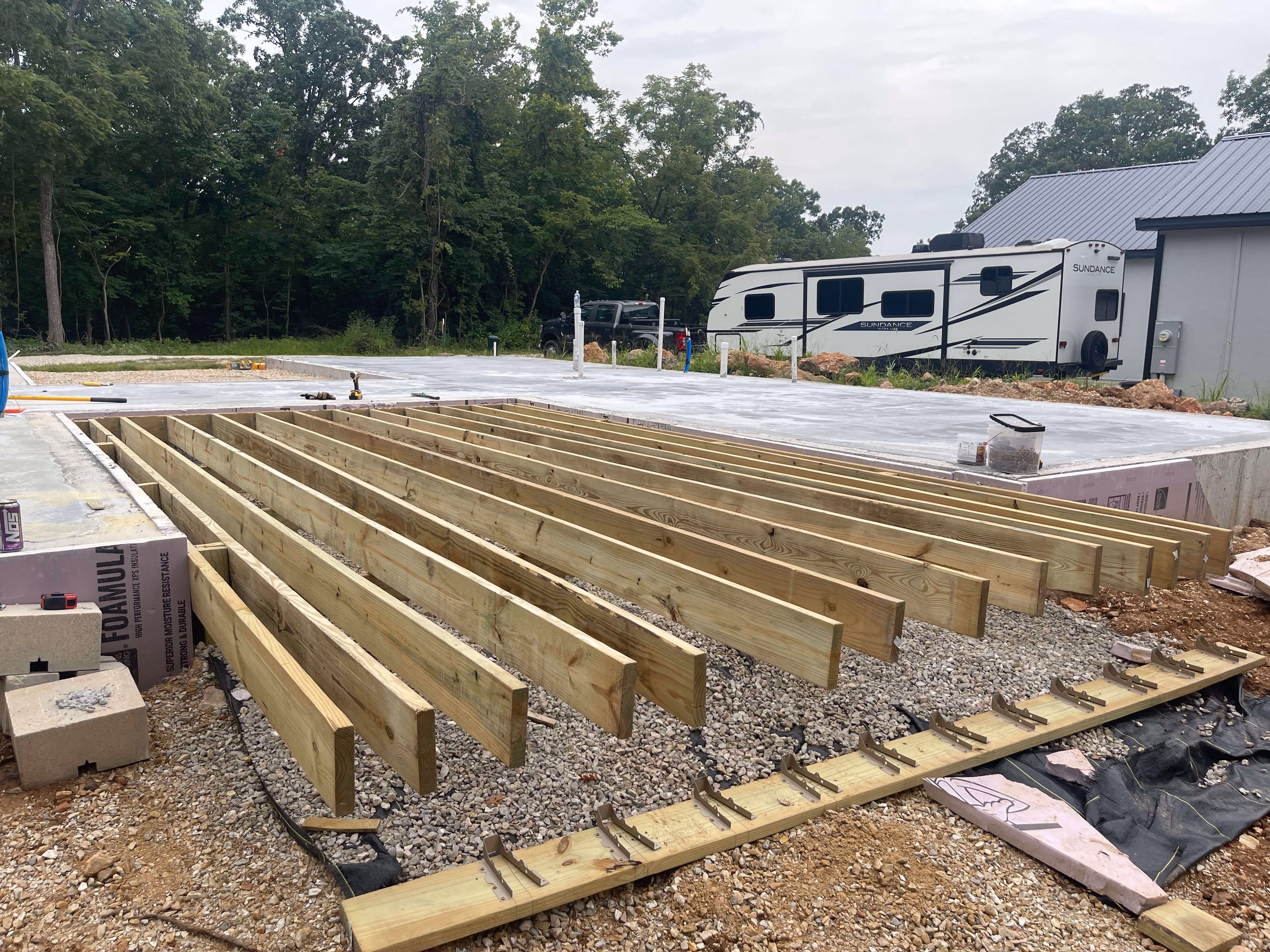 Wooden joists being installed on gravel foundation next to a concrete slab and an RV.