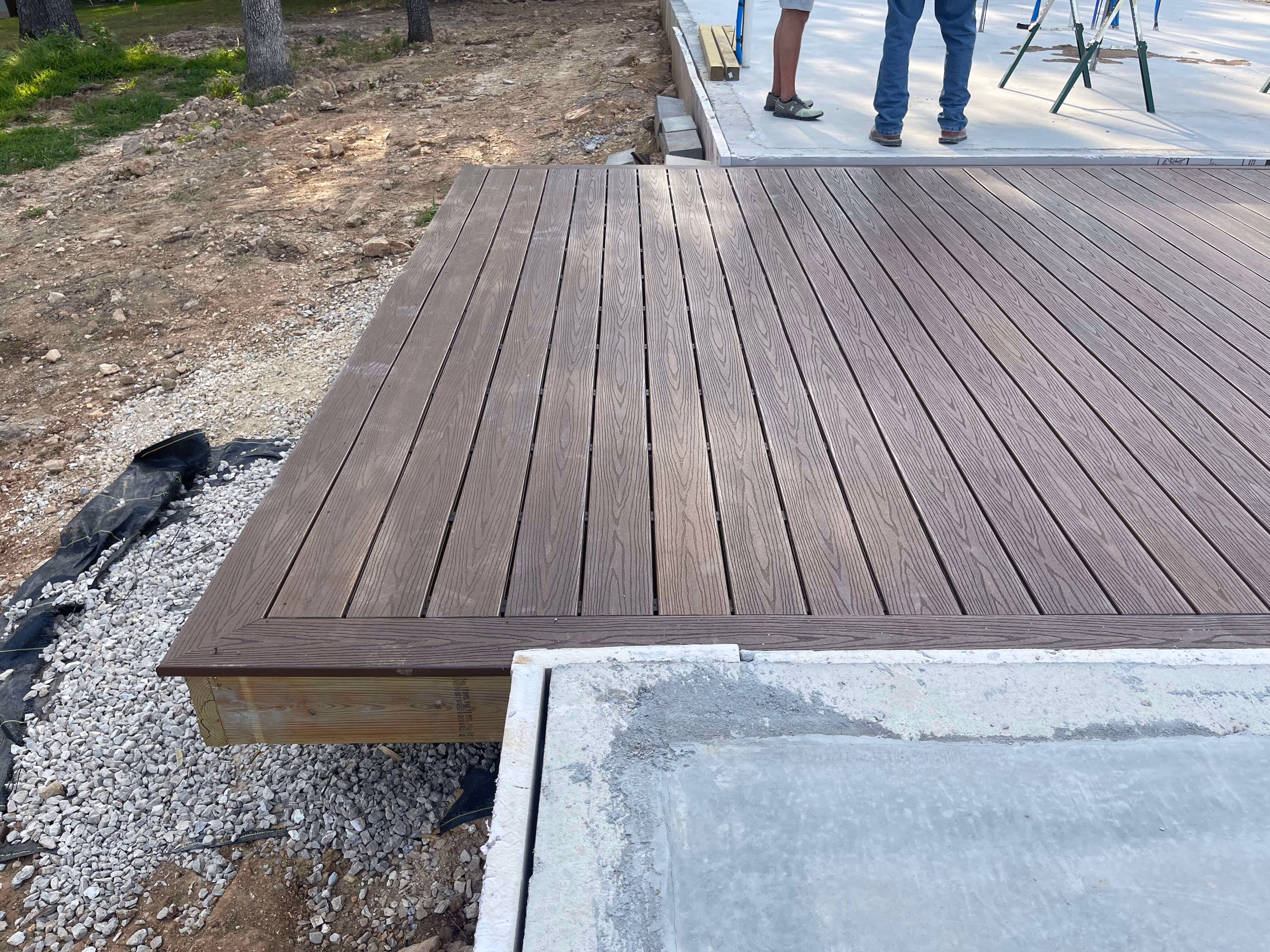 Brown composite wood deck next to concrete slab with two people standing in background.