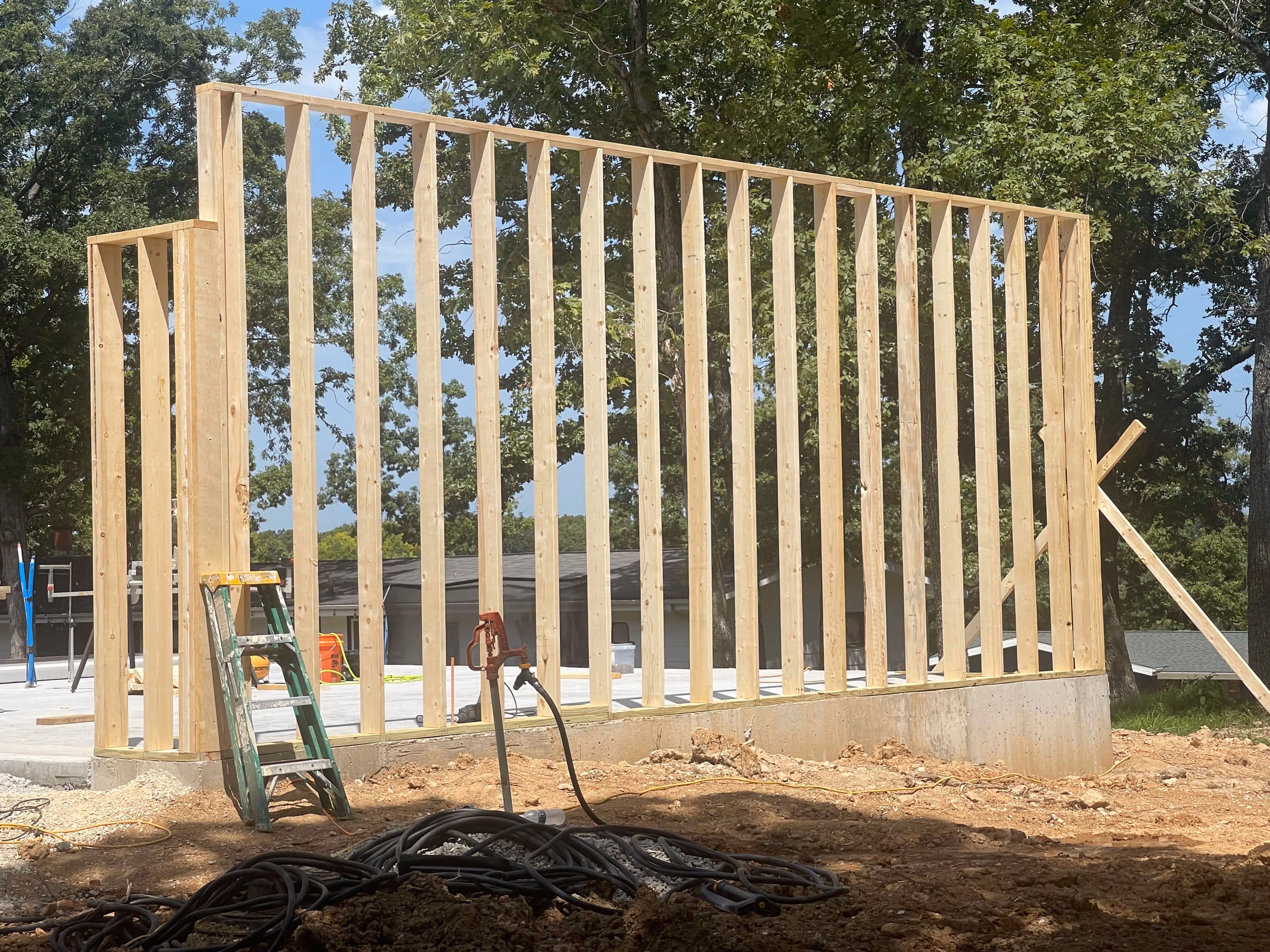 Wooden frame wall construction on concrete foundation outside with trees and construction tools.