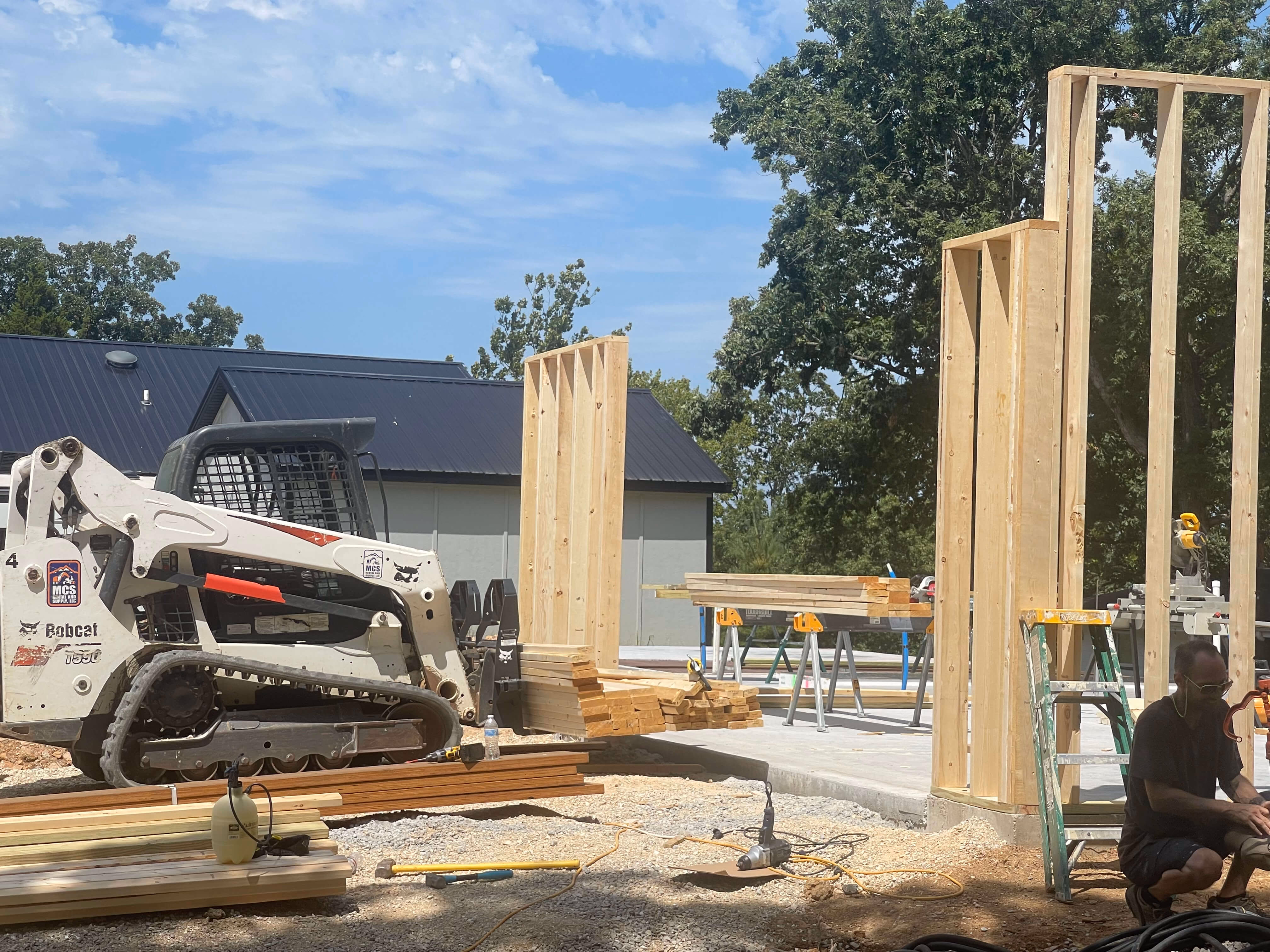Construction site with wooden framing, a Bobcat loader, stacked lumber, tools, and a man sitting near a ladder.