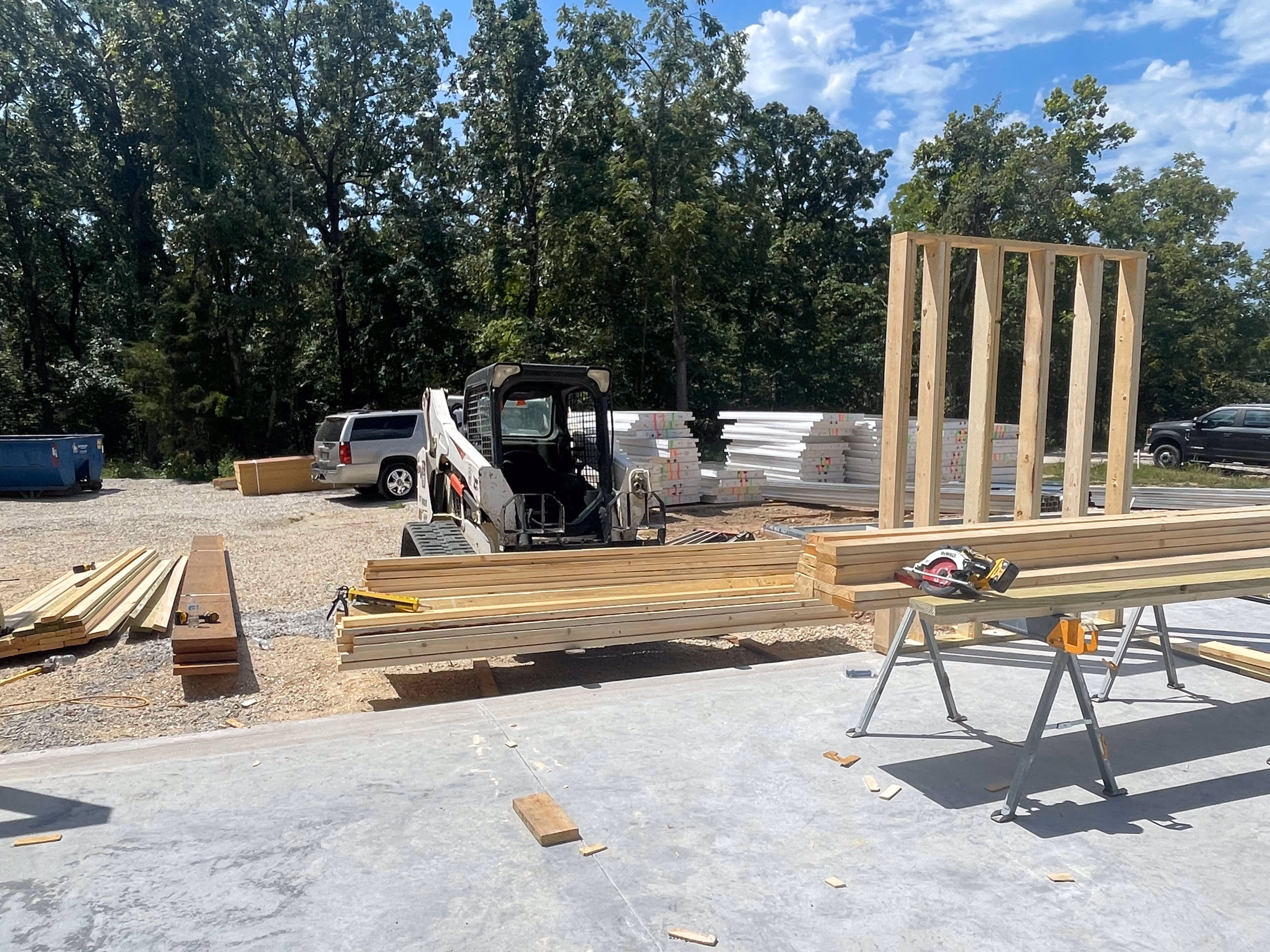 Construction site with piled lumber, a skid-steer loader, saw horses, and a partially framed wooden wall.
