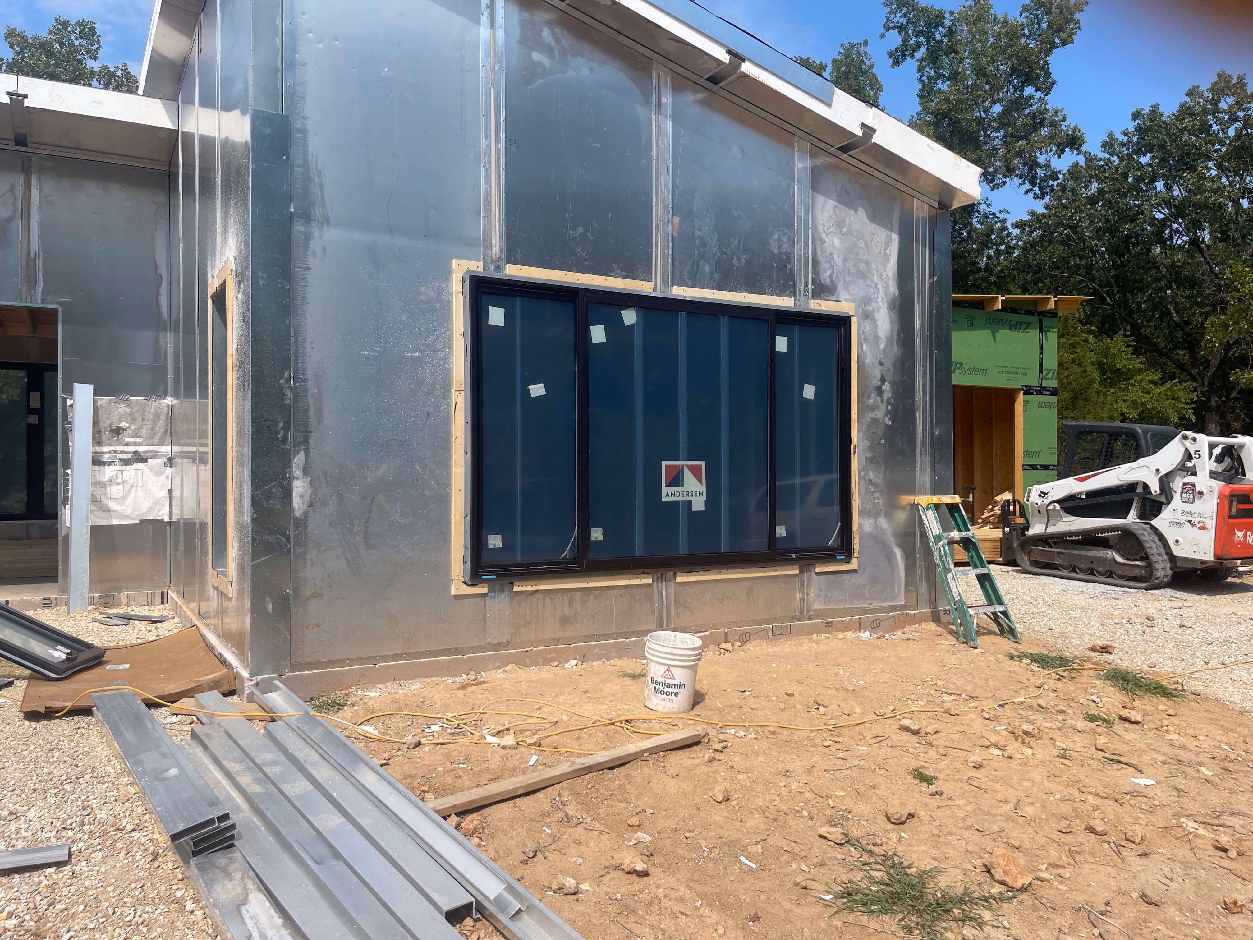 Construction site with metal-framed building, large Andersen window installed, ladder, and a skid steer loader.