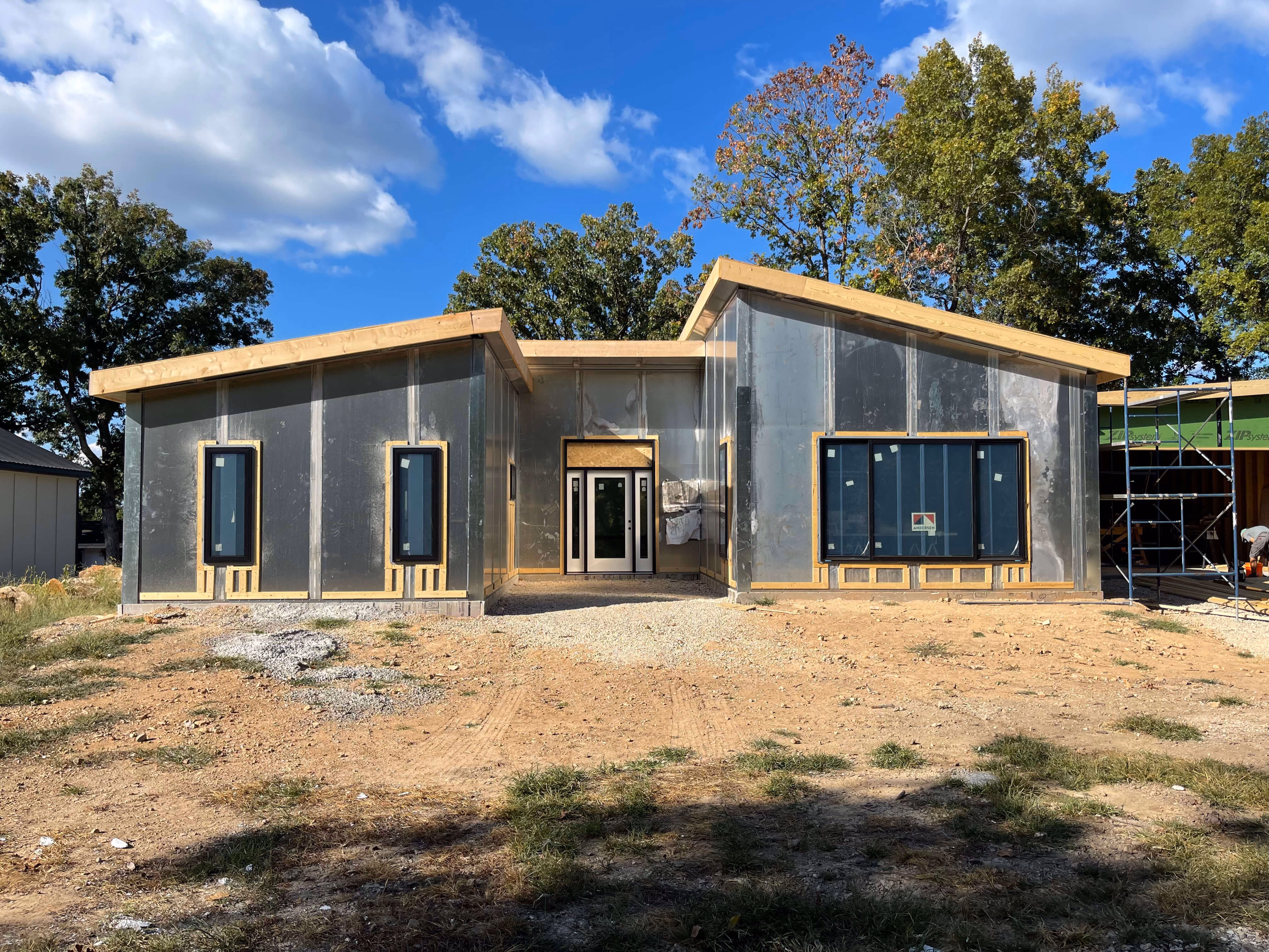 Modern house under construction with large windows, metal siding, and wood framing against blue sky.