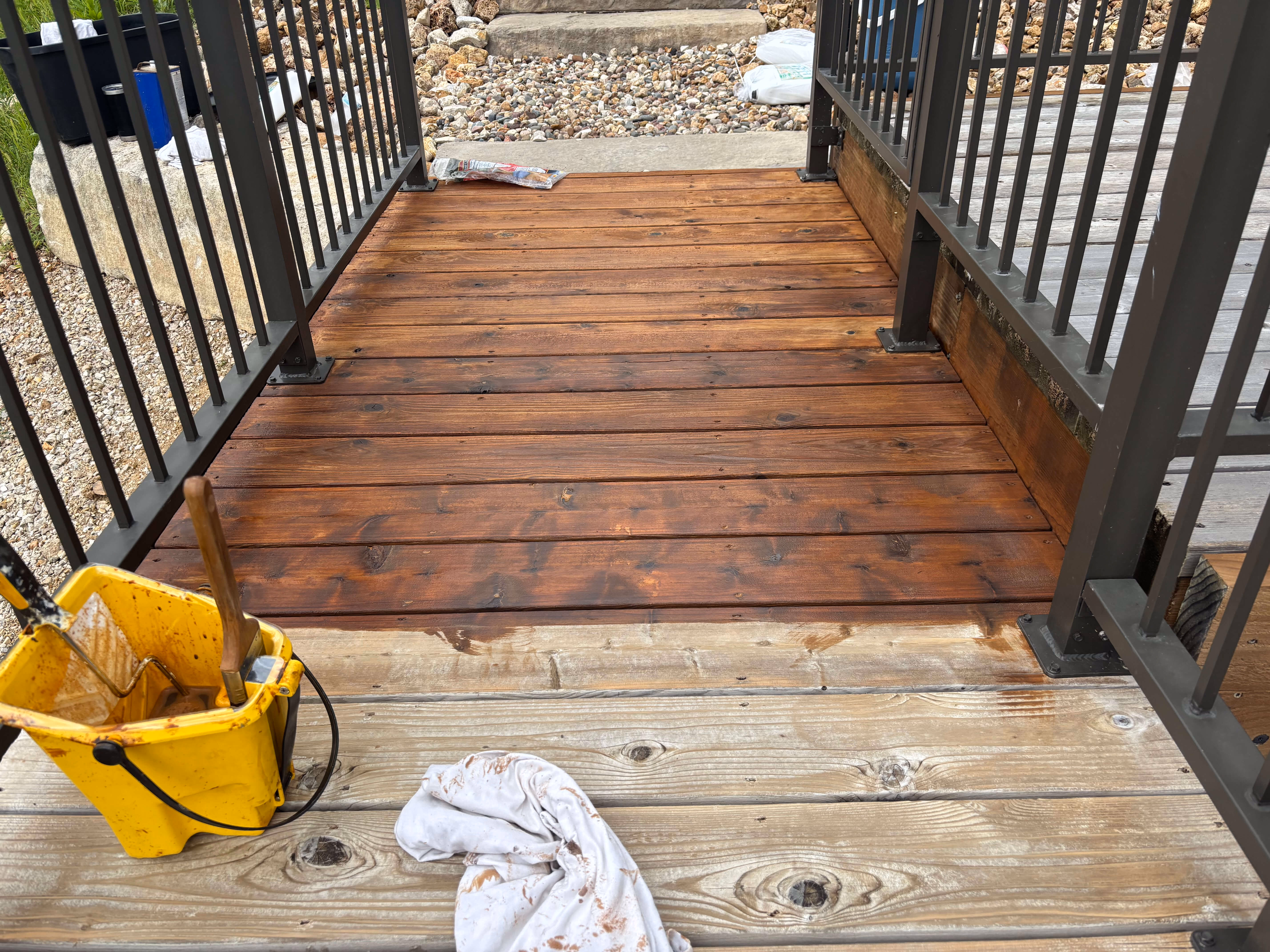 Partly stained wooden deck floor with metal railings and a yellow bucket with brushes.