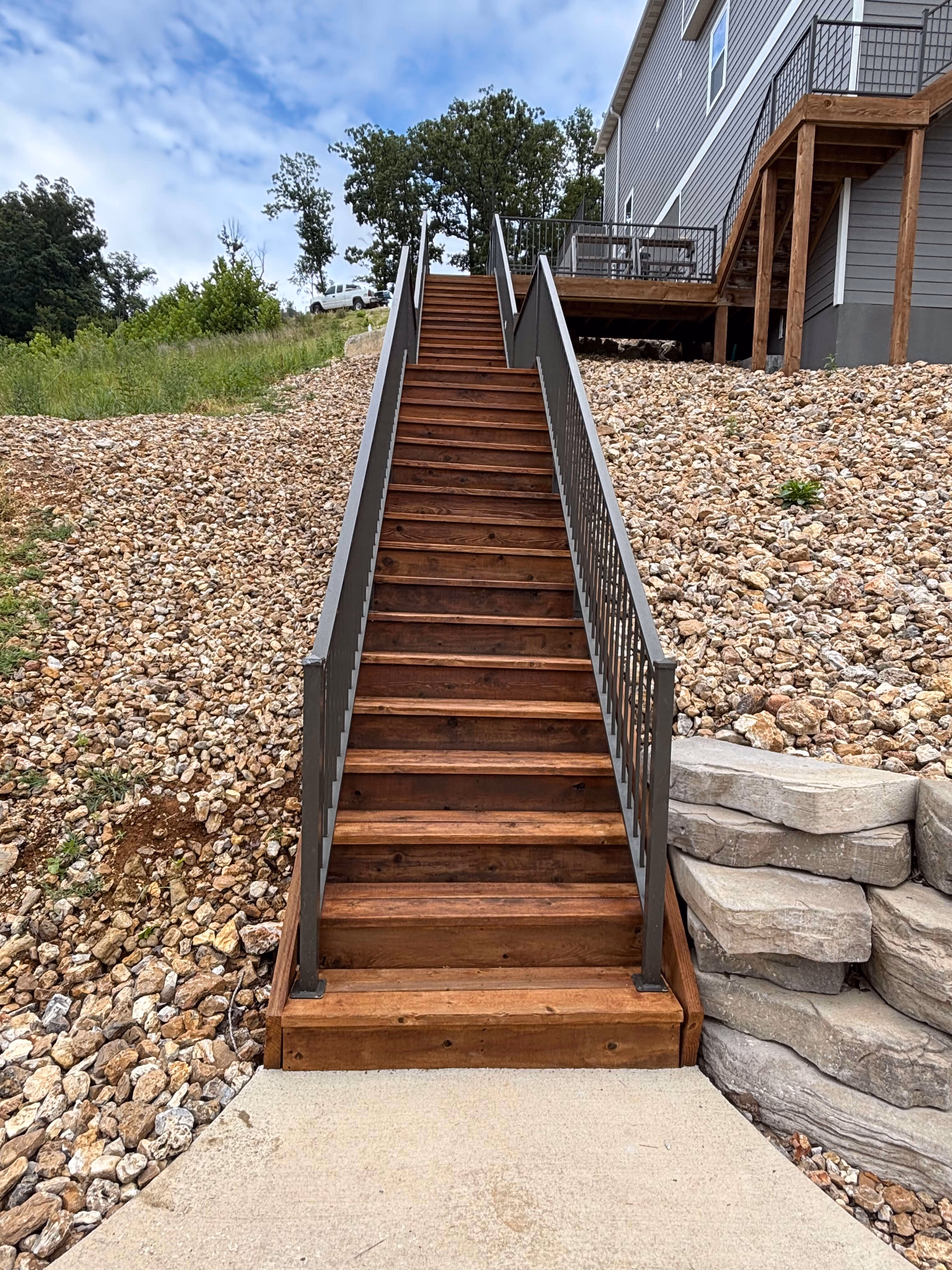 Wooden stairs with metal railings leading up a rocky slope to a house with a deck and patio furniture.
