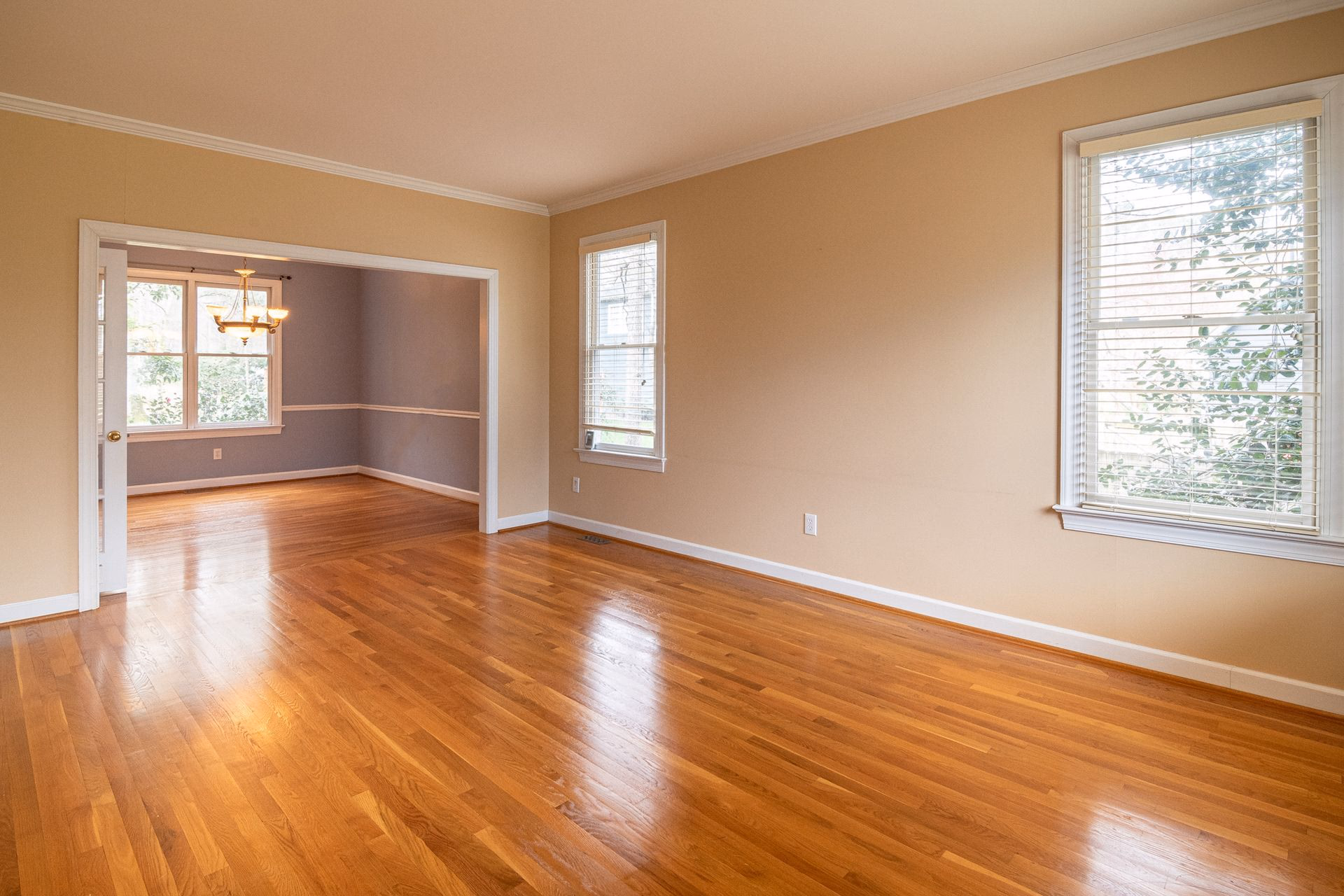 Empty room with polished hardwood floors, beige walls, and windows with white blinds.