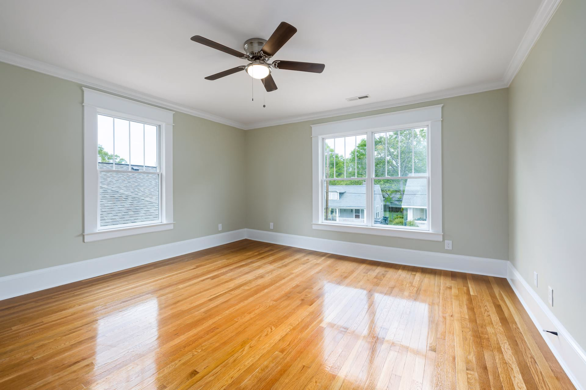 Empty room with shiny hardwood floors, light green walls, two large windows, and a ceiling fan with light.