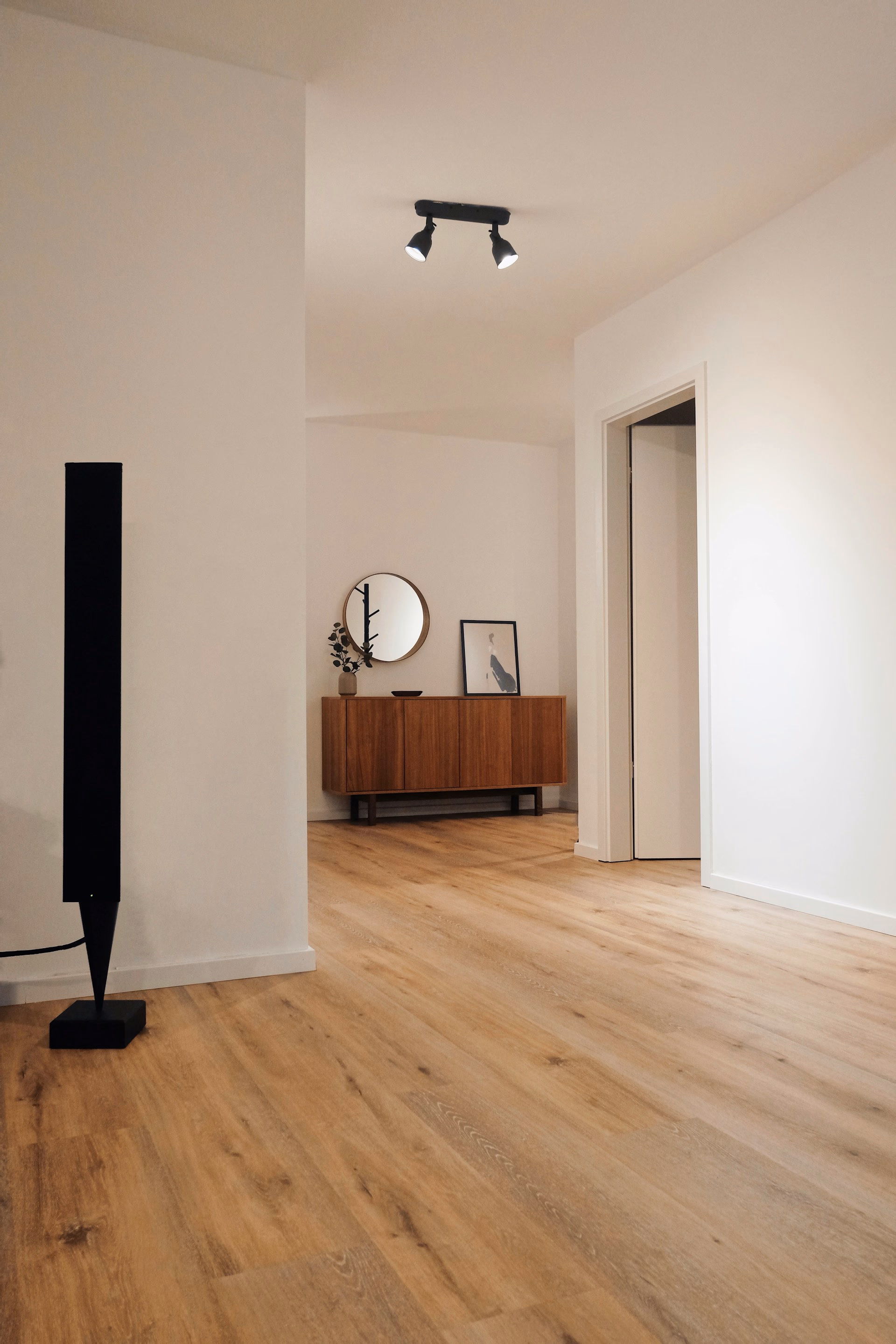Minimalist room with wooden floor, black floor lamp, wooden sideboard, round mirror, and framed art.